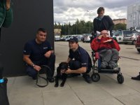Policjant kuca przy psie, obok mężczyzna w mundurze i niepełnosprawny na wózku inwalidzkim.